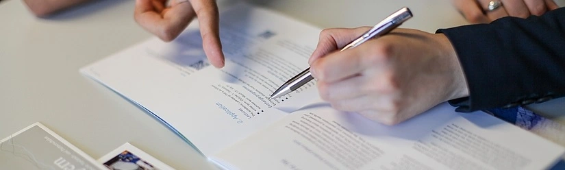 Ein Finger deutet auf Studienunterlagen, die Hand einer anderen Person hält einen Kugelschreiber