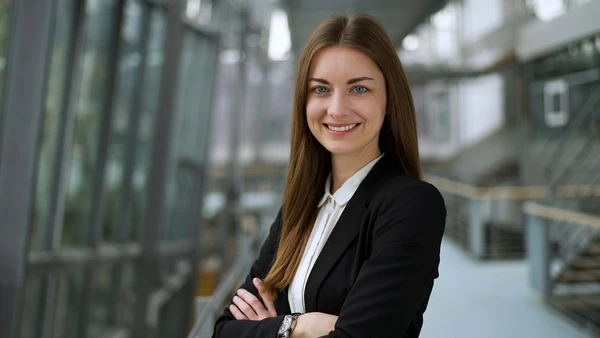 Eine junge Frau mit offenen langen braunen Haaren, gekleidet in weißer Bluse und schwarzem Blaser, im Hintergrund verschwommen das Treppenhaus der THI.