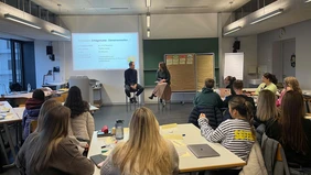 In einem Seminarraum, vorne an der Tafel sitzen zwei Erwachsene auf einem Stuhl und sprechen miteinander, hinten sitzen die Studierenden und hören dem Gespräch zu.