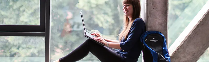 A student sitting in a window frame and working on a laptop