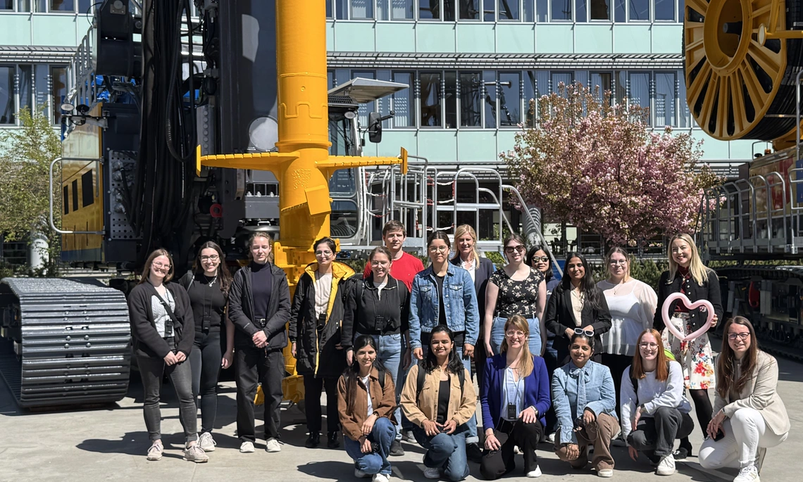 Teilnehmerinnen an der Exkursion bei der Bauer AG stehen zusammen für ein Gruppenbild vor einem Kran