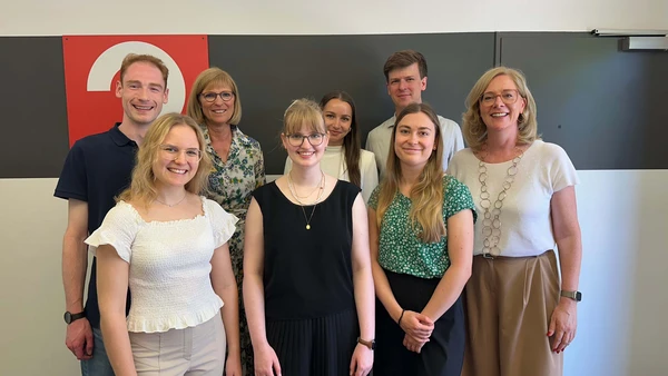 Ein Gruppenbild mit 8 Personen vor einer weißen Wand mit schwarzem Streifen. 
