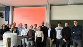 : Ein Gruppenfoto in einem Hörsaal, im Hintergrund die Präsentation und eine Tafel. 