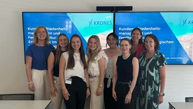Eine Gruppe von acht Frauen lächelt in die Kamera, im Hintergrund zwei Fernseher mit der Startseite der Präsentation.