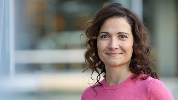 Eine Frau mit lockigen braunen Haaren und einem pinken Oberteil vor einem verschwommenen Hintergrund. 