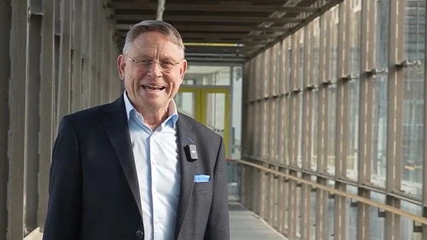 Ein Mann im Anzug in einem gläsernen Gang lächelt in die Kamera 