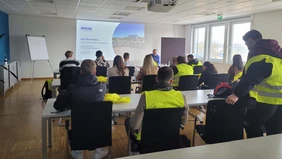 die Teilnehmer der Exkursion sitzen in einem Konferenzraum und schauen zur gezeigten Präsentation, einige Teilnehmer tragen gelbe Sicherheitswesten